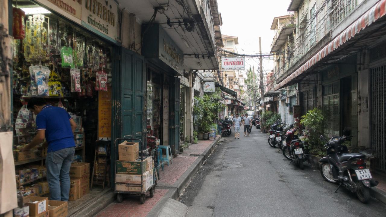 ภาพจำลองซอยตึกแถวเก่าย่านเมืองเก่ากรุงเทพฯ มีร้านค้าขนาดเล็กและเจ้าของร้านจัดของ ไม่ใช่สถานที่ของลูกค้าจริง (Illustrative old shophouse lane in Bangkok old town with small shops and a shopkeeper arranging goods, not an actual client location)